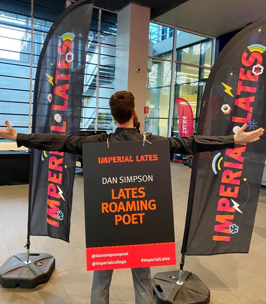 Dan, arms outstretched, wearing a sandwich board reading 'Imperial Lates Roaming Poet'