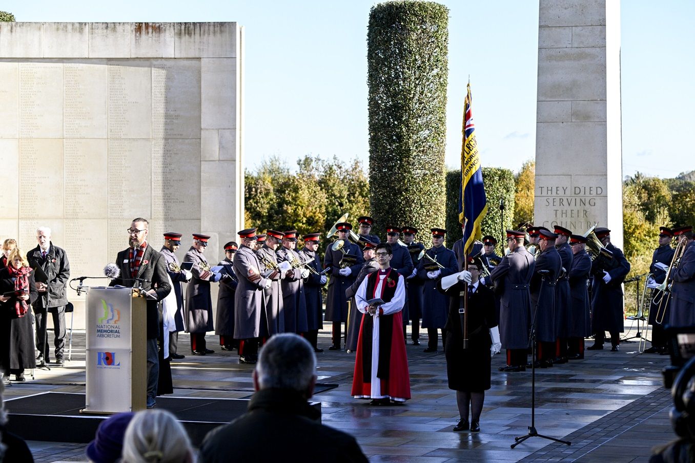Dan reading at the lectern during a Remembrance service at the National Memorial Arboretum, with a military band behind